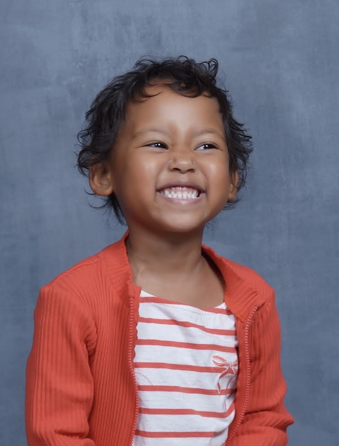 Sofia school picture of pre-K black girl with short curly hair and a big smile wearing and orange and white striped top under an orange sweater with a zipper