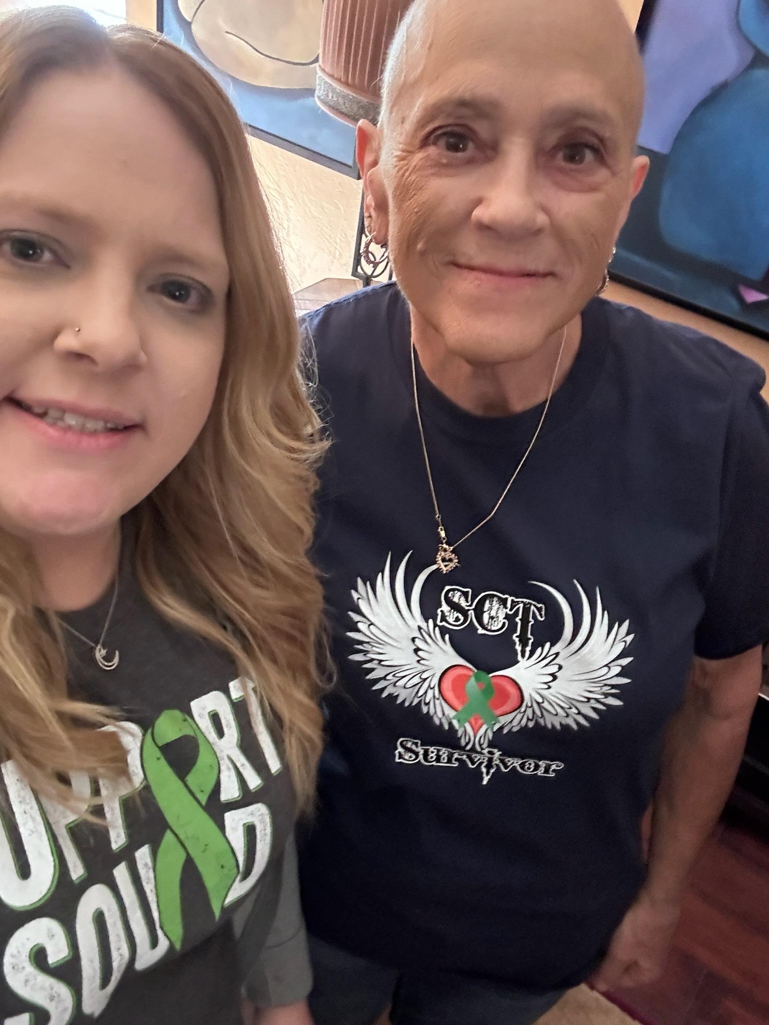 Susanne middle aged white woman with a bald head wearing hoop earrings and a heart necklace and a SCT Survivor t-shirt standing next to a young woman with a nose ring and Support Squad t-shirt