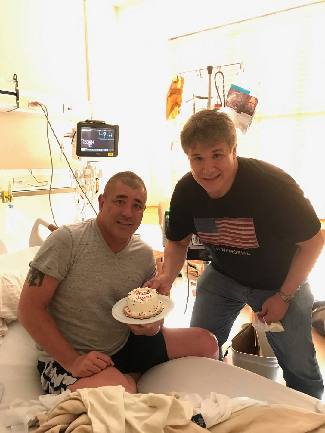 Greg two middle aged white men, one sitting on a hospital bed wearing a grey t-shirt and black shorts the other wearing a flag shirt and jeans both holding a cake