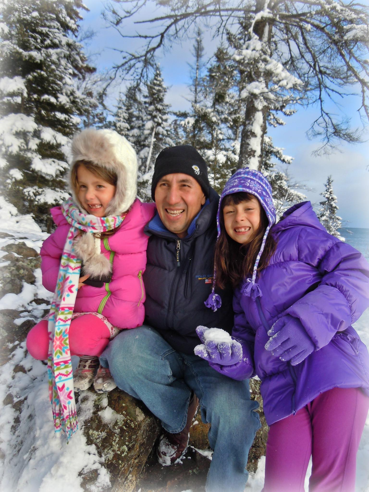 Joaquin middle aged hispanic man wearing navy beanie and ski jacket with two hispanic girls one in a pink jacket and one in a purple jacket holding a snowball in front of snow covered trees