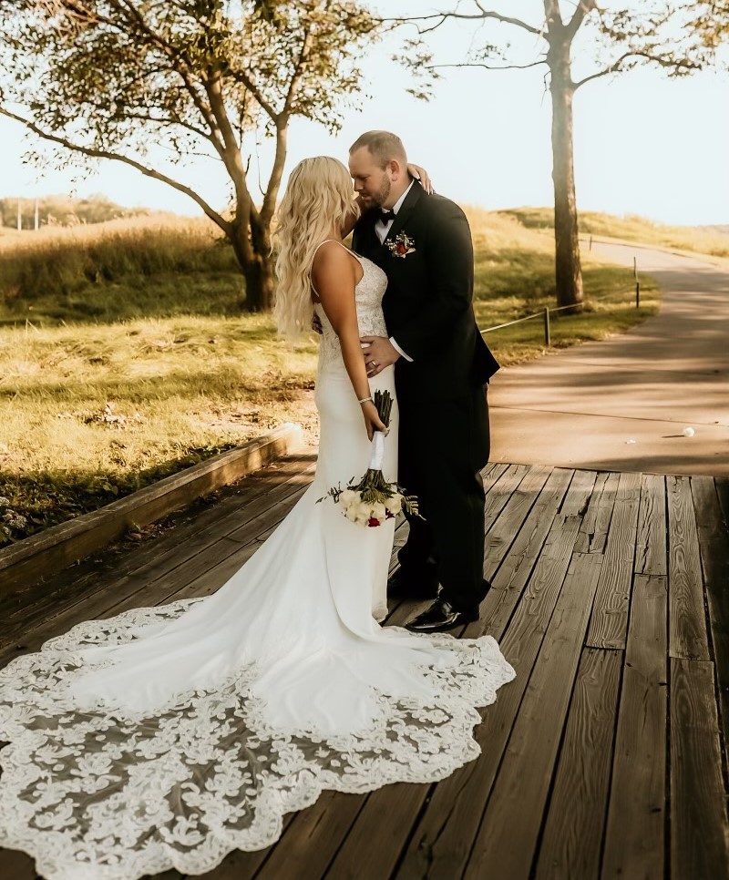 Kayla young woman with long blond hair in a white spagetti strap wedding dress holding a bouquet with arm around a young white man with beard and mustache.