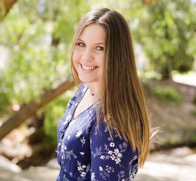 Maddie young white female with long brown hair and a big smile wearing a blue and white flowered dress standing in front of a blurred background of trees