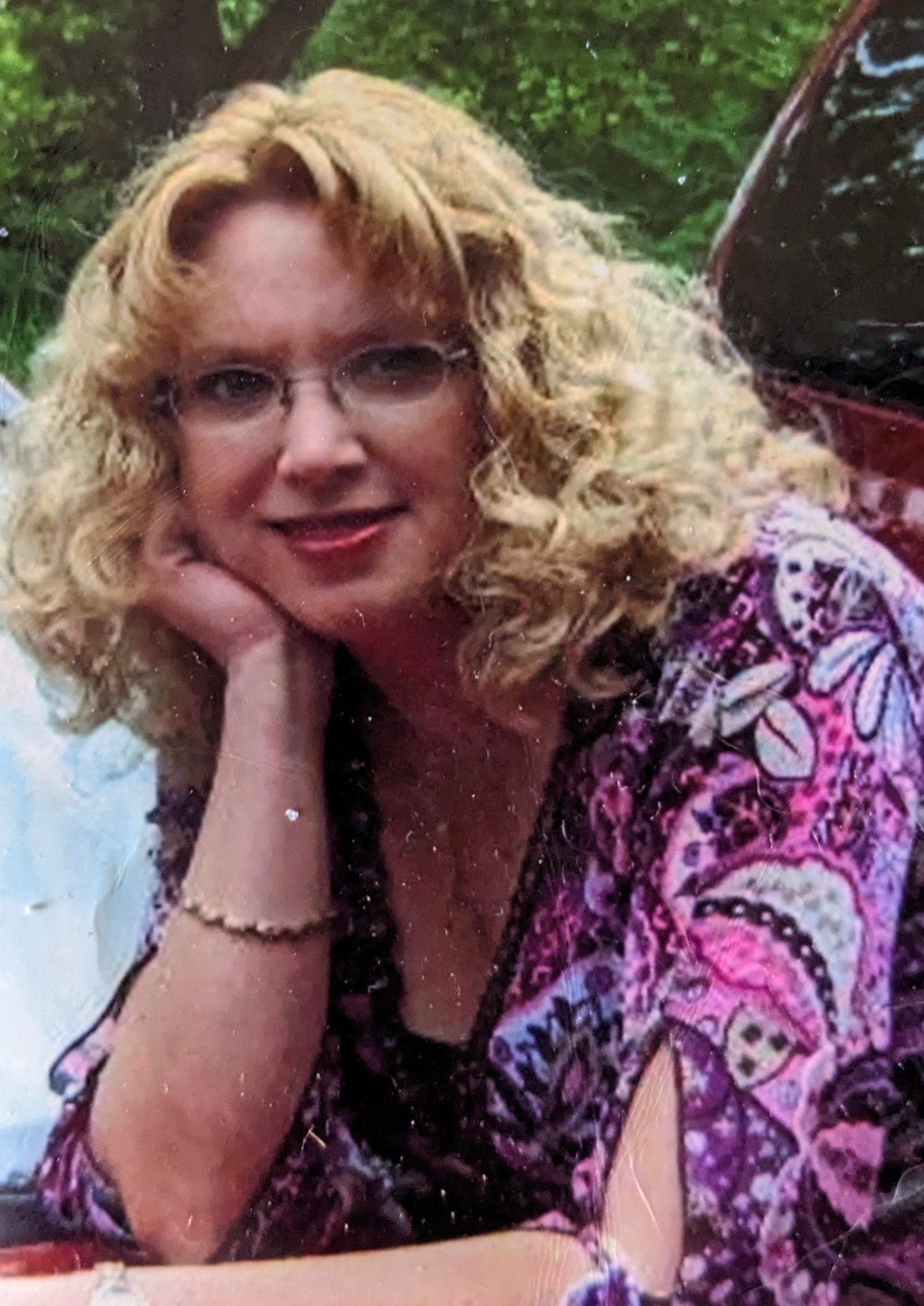 middle aged white woman with curly blonde hair and glasses with her chin resting on her hand wearing a pink and purple print shirt