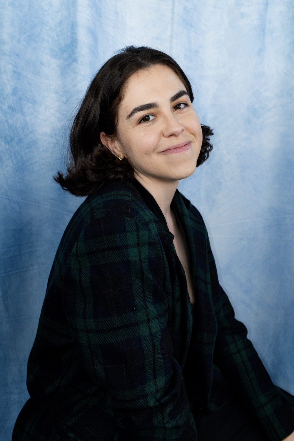 Crystal young hispanic woman with dark hair and gold hoop earrings smiling and wearing a blue and plaid top with blue photo background