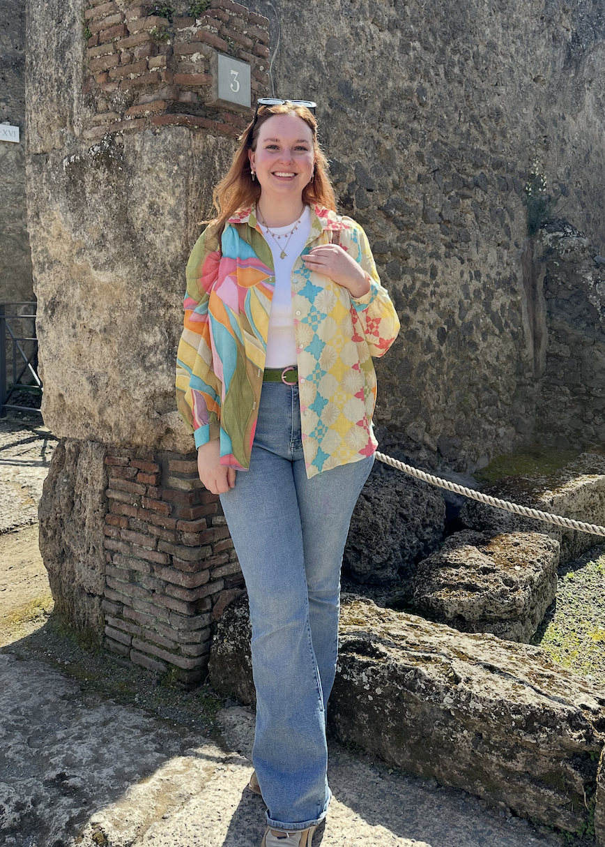 Eva young white woman with reddish hair sunglasses on her head in a white tshirt under a multi color shirt with blue jeans standing in front of a stone wall.