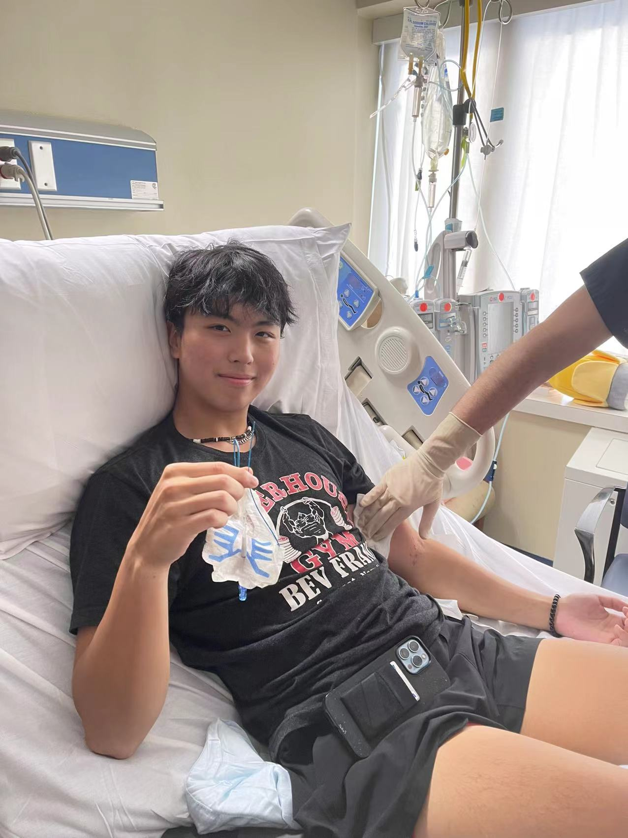 Kevin young asian young man with black hair wearing a puka shell necklace and black gym tshirt and shorts while laying in a hospital bed