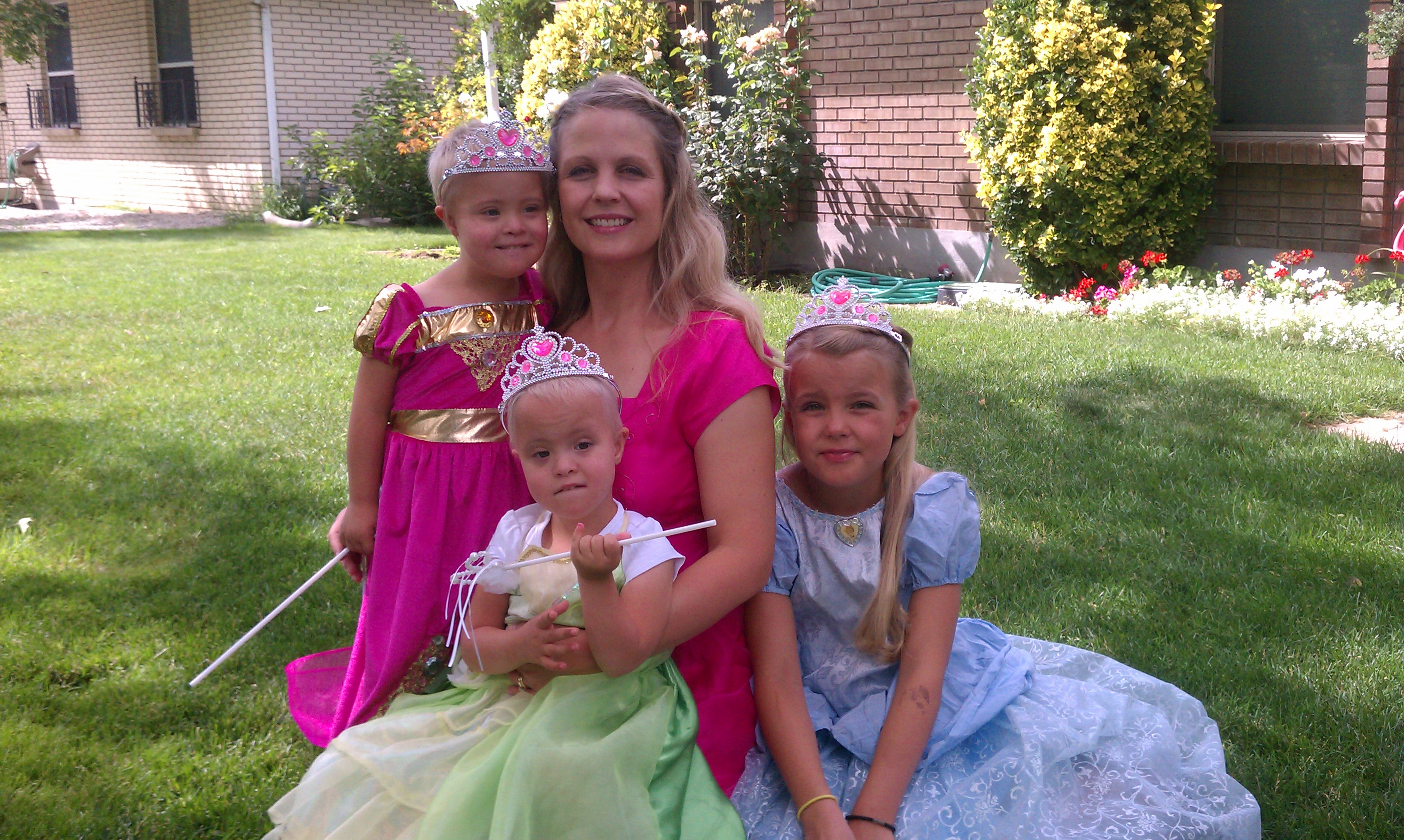 Kim young white mother with long blond hair in a pink dress with three little girls wearing princess outfits and crowns sitting outside