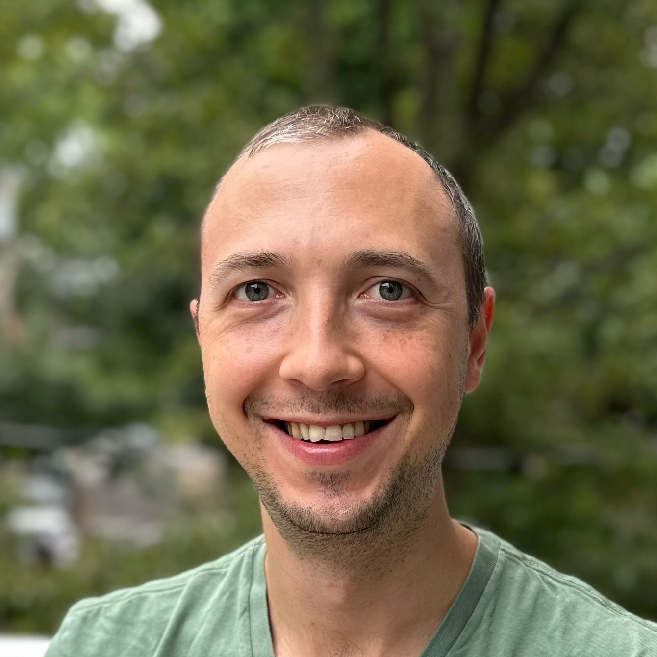 Robert, young white male with receeding hairline and a stubble mustache and beard wearing a green tshirt standing in front of blurred trees