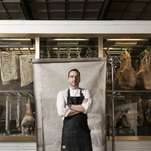 Steve young white man with short hair glasses and a beard and mustache wearing a white chef's coat under a denim apron standing in front of a canvas that is front of windows showing meats being cured