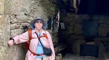 Kendra stands against a stone wall, wearing a pink shirt, hiking gear, and a hat.