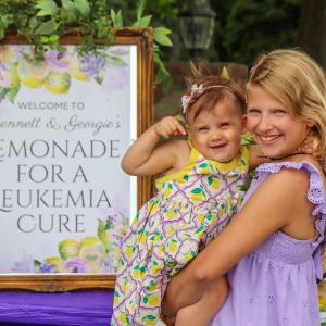 Blond girl in purple dress holding younger girl by lemonade stand sign.