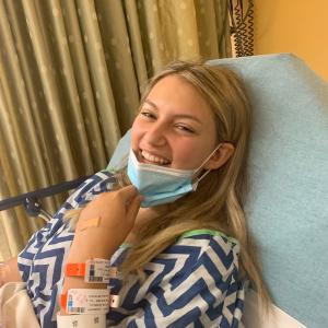 Blonde teen girl with leukemia in blue and white medical gown pulling down a face mask