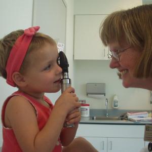 Little girl with red headband and shirt holding medical instrument next to a woman with glasses