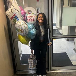 Smiling black teen girl with lymphoma in black jacket and pants holding mylar balloons and a gift bag