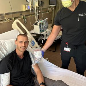Young man in black t-shirt with leukemia in a hospital bed with a medical professional standing next to him wearing a mask