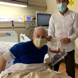 Man in blue shirt and mask in a hospital bed with a medical worker holding a bag of stem cells