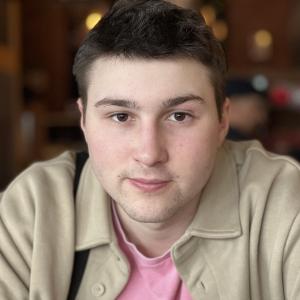 young white man with dark brown hair and chin stubble wearing a pink shirt and beige jacket