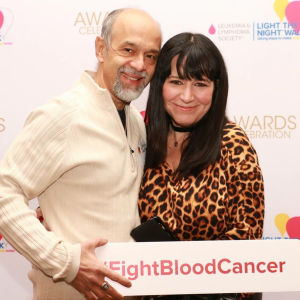 an older hispanic couple hugging in front of LTN wall he in a cream shirt holding a sign that says FightBloodCancer and her in an animal print top