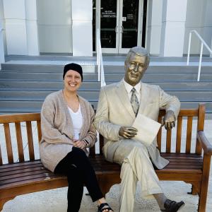 bald white woman wearing a knit cap tan sweater and white shirt sitting on a bench next to a statue