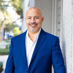 balding middle aged hispanic man with graying beard and mustache smiling and wearing a royal blue jacket and white shirt