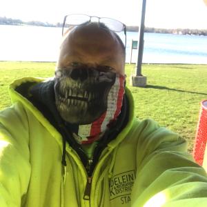 balding white man with sunglasses on his head wearing a monster mask and yellow hoodie in front of water