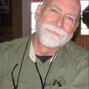 older white gentleman with white beard and mustache wearing a long-sleeved green shirt with a pair of glasses hanging around his neck