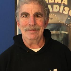 older white man with bushy eyebrows and a mustache wearing a black hoodie that says Italia standing in front of a movie poster