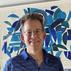 older white man with spiky hair wearing glasses and a blue striped shirt in front of mosaic artwork