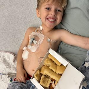 smiling young white boy laying in bed with a port in with a box of rolls