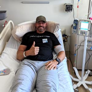 middle aged white man in a ball cap with a scruffy beard and mustached wearing a black t-shirt lying in a hospital bed giving a thumbs up