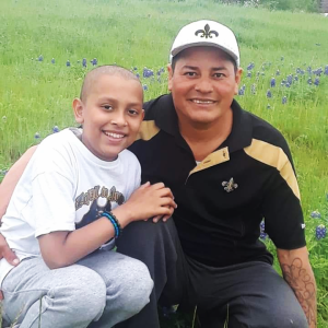 young hispanic child with little hair in white t-shirt and sweat pants with his father who is wearing a New Orleans Saints hat and a black and gold shirt, both in a field of bluebonnets