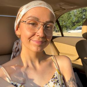 young white woman sitting in a car wearing a head scarf, wire rim glasses, a floral sundress witha nose ring