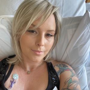 young woman with blond hair and long eyelashes lying in hospital bed wearing a black sleeveless top showing a port and a tattoo