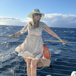 middle aged white woman wearing a straw hat sunglasses and a floral dress leaning on a boat's railing