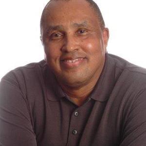 older black man smiling wearing a burgundy colored shirt with his arms crossed