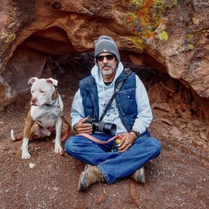 older white man with beard and mustache wearing a knit cap and puffy blue vest holding a camera sitting by his dog