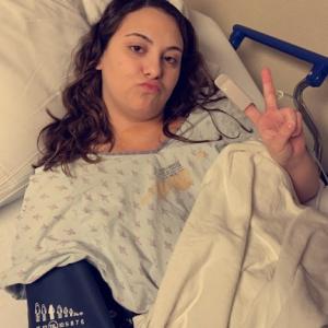 young woman with long brown hair wearing hosptial gown puckering her mouth and giving a peace sign lying in a hospital bed