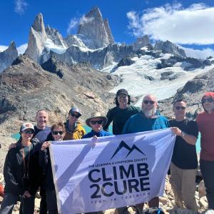 a group of nine people standing at the top of a mountain holding a Climb2Cure banner