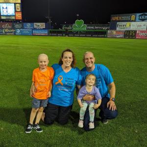 little white boy who is bald wearing an orange t-shirt and blue shorts next to his mom sister and dad wearing blue and orange tshirts on a ball field