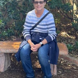 middle aged white woman short brown hair sitting on a bench and wearing a blue striped shirt and jeans with a jacket tied around her waist