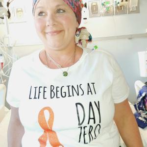 middle aged white woman wearing a colorful headscarf and white t-shirt with an orange ribbon on it sitting on a hospital bed