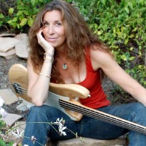 middle aged white woman with long brown hair wearing and orange tank and jeans with a guitar on her lap sitting in a garden