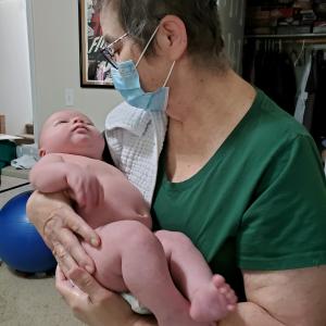 middle aged white woman with short hair and glasses wearing a face mask and a green shirt holding a baby