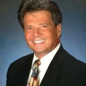 older white man headshot smiling wearing a brown suite white shirt and brown and blue tie