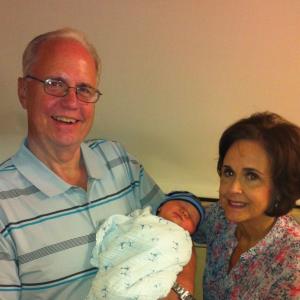 older white man with glasses and blue striped shirt holding a baby with an older woman with dark hair and a floral blouse