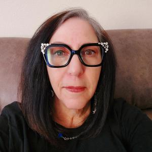 older white woman with dark long hair wearing black chunky glasses with bling on them and a black top