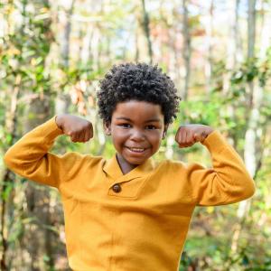 smiling young black boy standing in front of trees wearing a gold sweater and smiling and making muscle man arms