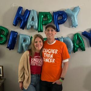 young white couple standing in front of a birthday balloon sign she has blond hair and burgundy t-shirt on he is wearing a ball cap and orange t-shirt