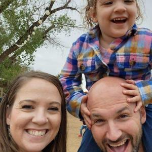 young white couple with their daughter outside little girl has on plaid shirt dad is bald with a mustache and beard mom has long brown hair