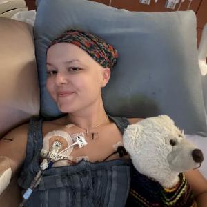 young white woman wearing a head scarf and gray top lying in a hospital bed with a stuff white bear